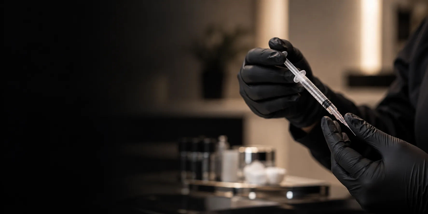 Black-gloved hands preparing a syringe in a dimly lit treatment room, fading into black on the left
