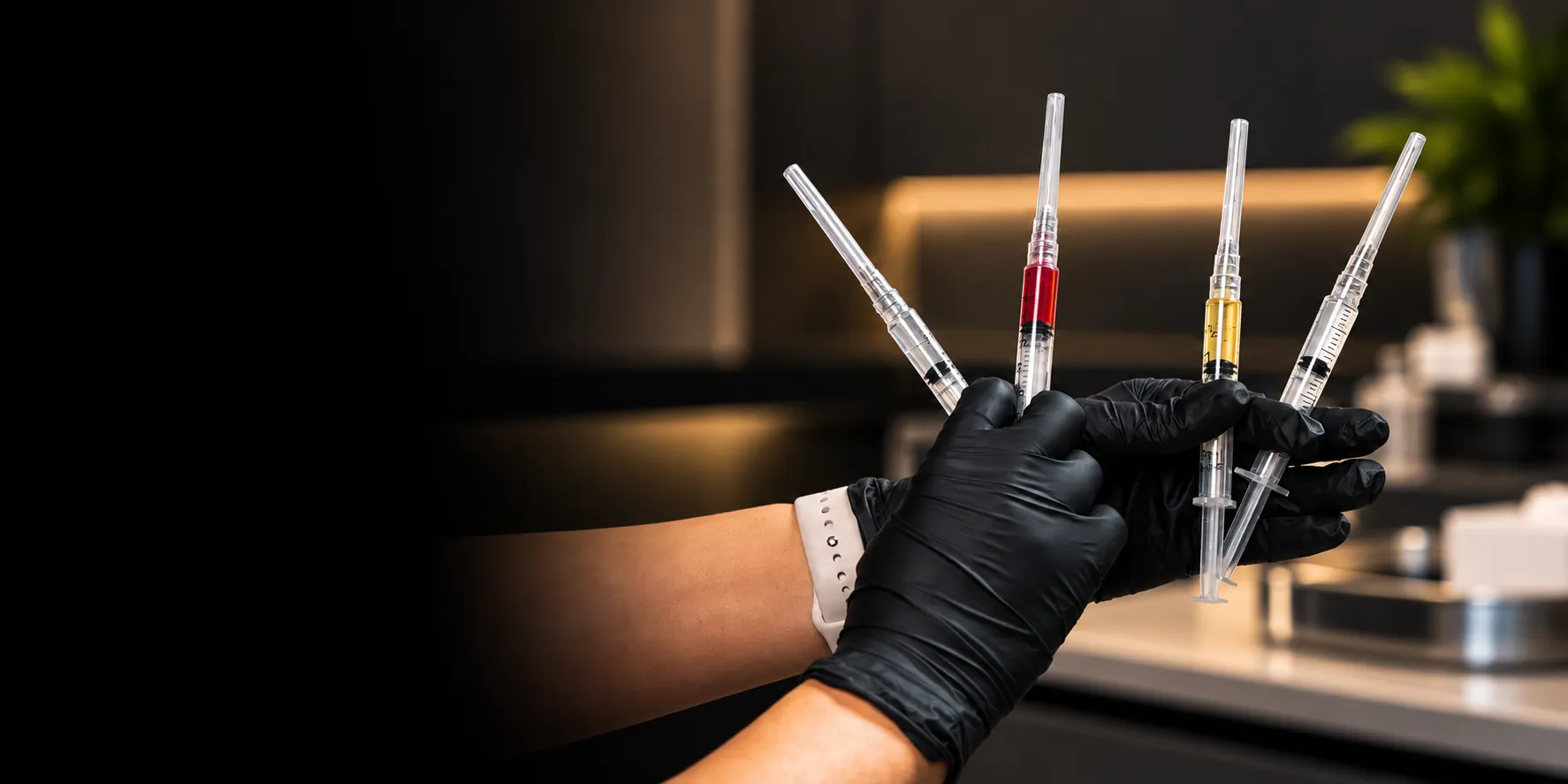 Black-gloved hands fanning four syringes, one filled with red liquid and one with yellow, in a dim treatment room, fading into black on the left